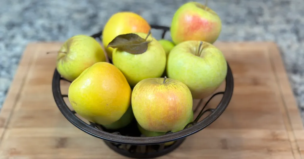 A metal basket full of apples that are yellow and green with a touch of red sitting on a cutting board on the counter.