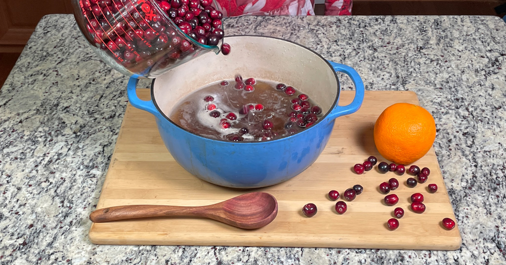 Cranberries being added to a blue enameled dutch oven containing a boiling sugar and water mixture.