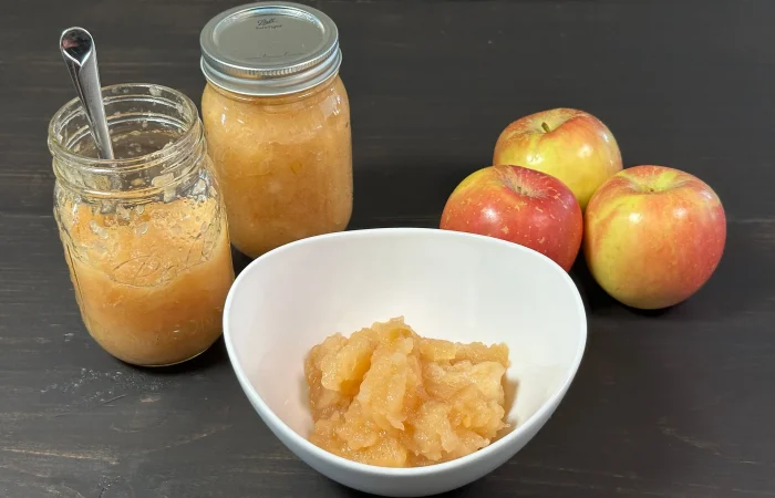 Chunky applesauce in a white bowl. Two mason jars of water bath canned apple sauce and three yellow and red apples sitting on a black background.