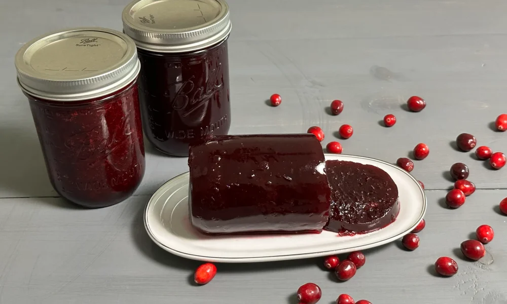 Cranberry Jelly sitting on a plate that has been sliced. Two mason jars of cranberry jelly on the side of the plate that were water bath canned.