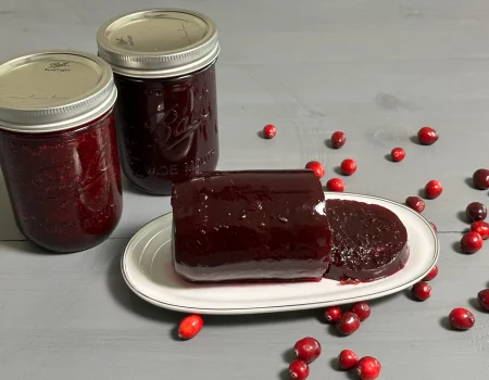 Cranberry Jelly sitting on a plate that has been sliced. Two mason jars of cranberry jelly on the side of the plate that were water bath canned.