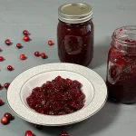Cranberry sauce in a bowl with an open canning jar of cranberry sauce with a spoon.