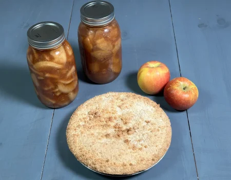 Two quart sized mason jars of Apple Pie Filling that have been water bath canned. Apple pie made with apple pie filling with two red and yellow apples sitting on a blue background.