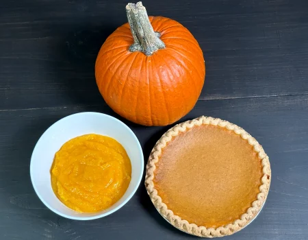 Pumpkin puree in a white bowl with a pumpkin and a pumpkin pie sitting on a dark background.