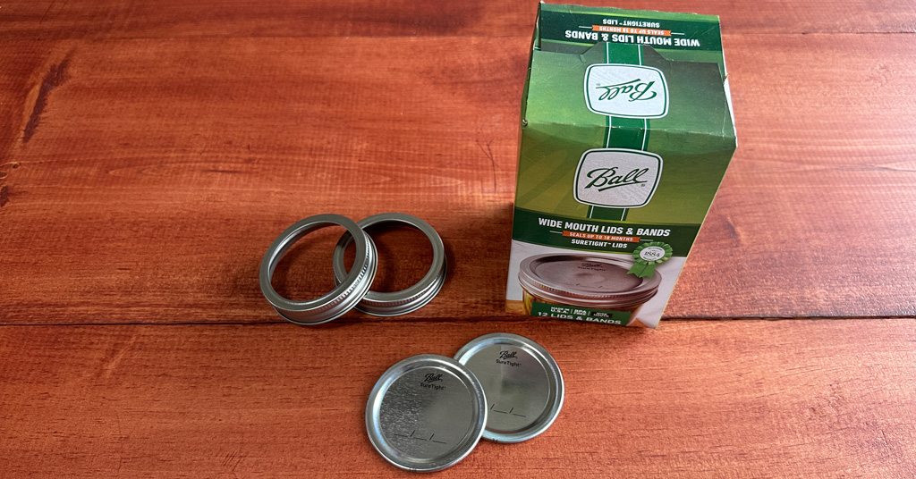 Box of Ball mason jar lids and rings with two rings and two lids sitting on a counter.