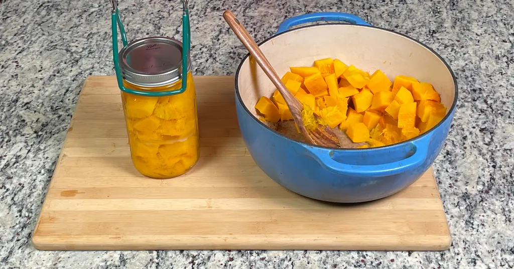 Jar lifter lifting quart sized mason jar of pumpkin cubes to be put in the pressure canner. Blue enameled dutch oven with pumpkin cubes waiting to be put into mason jars.