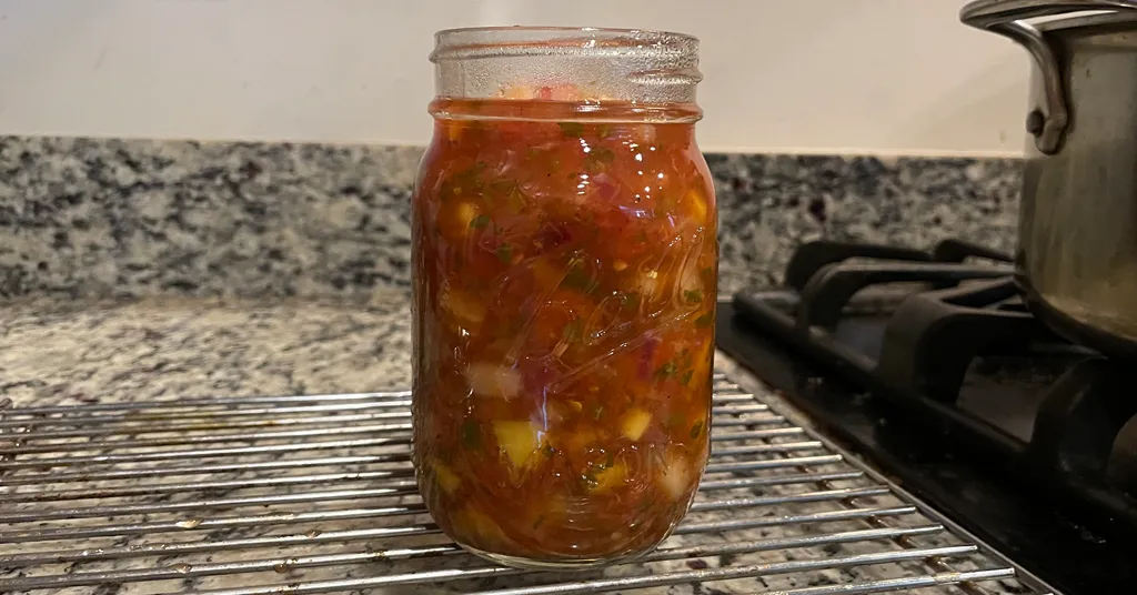Mason jar filled with salsa just before being water bath canned, sitting on a metal cooling rack.