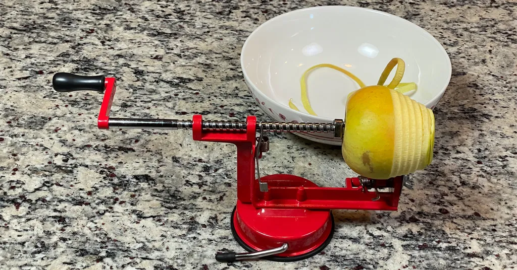 A red Apple Peeler, Corer, and Slicer. Peeling an apple with the peel going into a bowl.