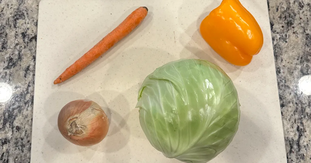 A head of green cabbage, one carrot, one orange pepper, and one onion sitting on a white cutting board on a counter.