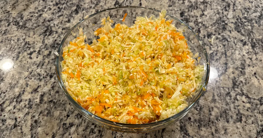 Shredded cabbage, carrots, peppers, and onions mixed together in a glass bowl on the counter to make coleslaw.