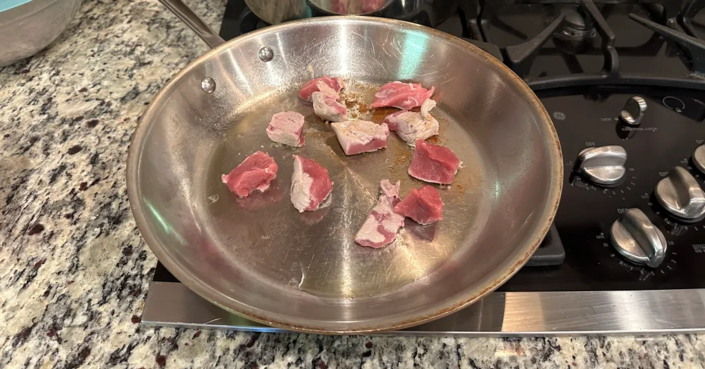 Pieces of pork tenderloin being browned in a stainless steel frying pan.