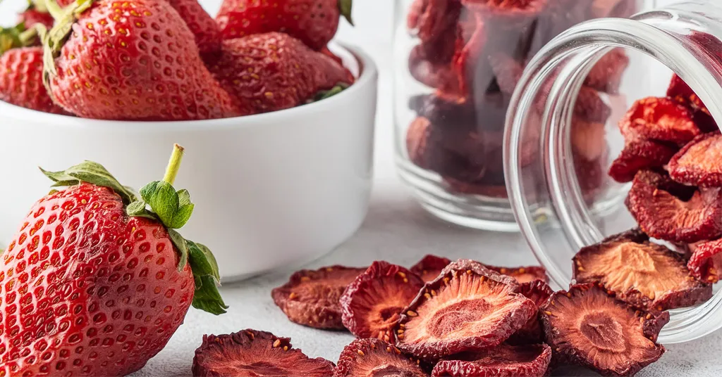 Some dehydrated strawberry slices spilling out of a glass jar sitting next to a white bowl of red ripe strawberries.