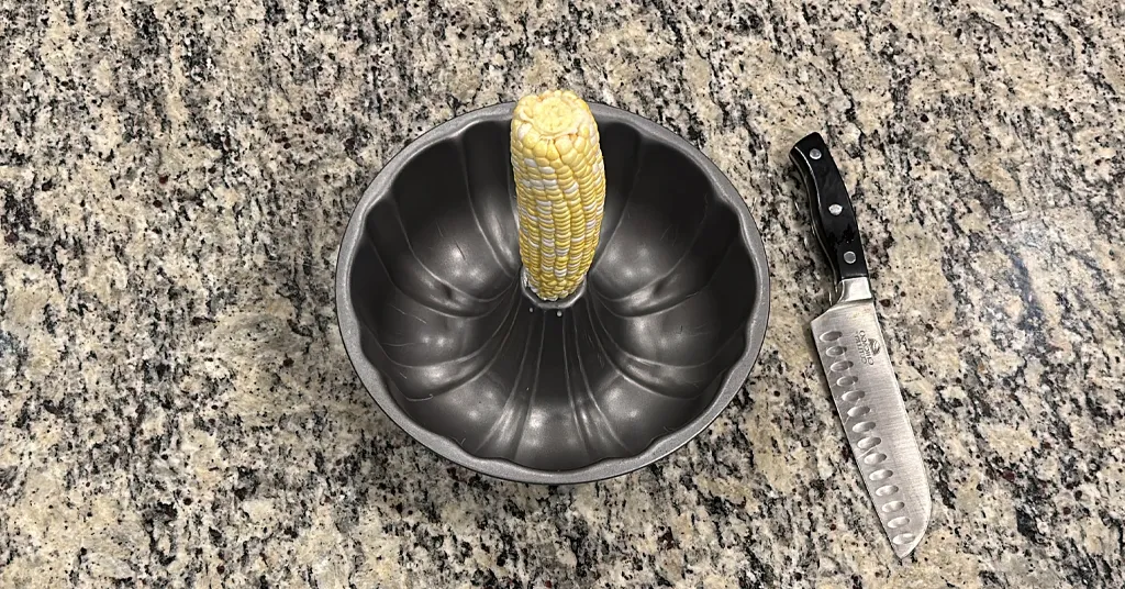 A fresh ear of corn positioned upright in the center of a bundt pan, with a large kitchen knife lying next to it on a granite countertop.