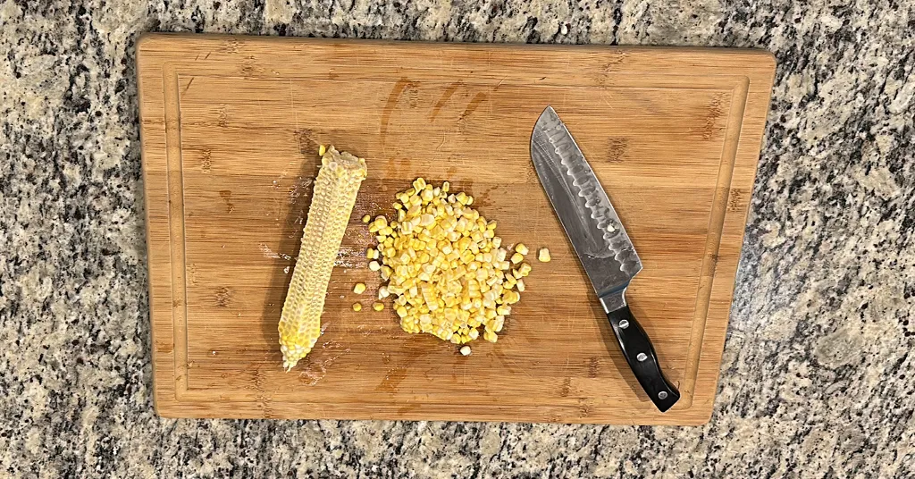 A wooden cutting board with a pile of freshly cut corn kernels, an empty corn cob, and a large kitchen knife placed on a granite countertop.