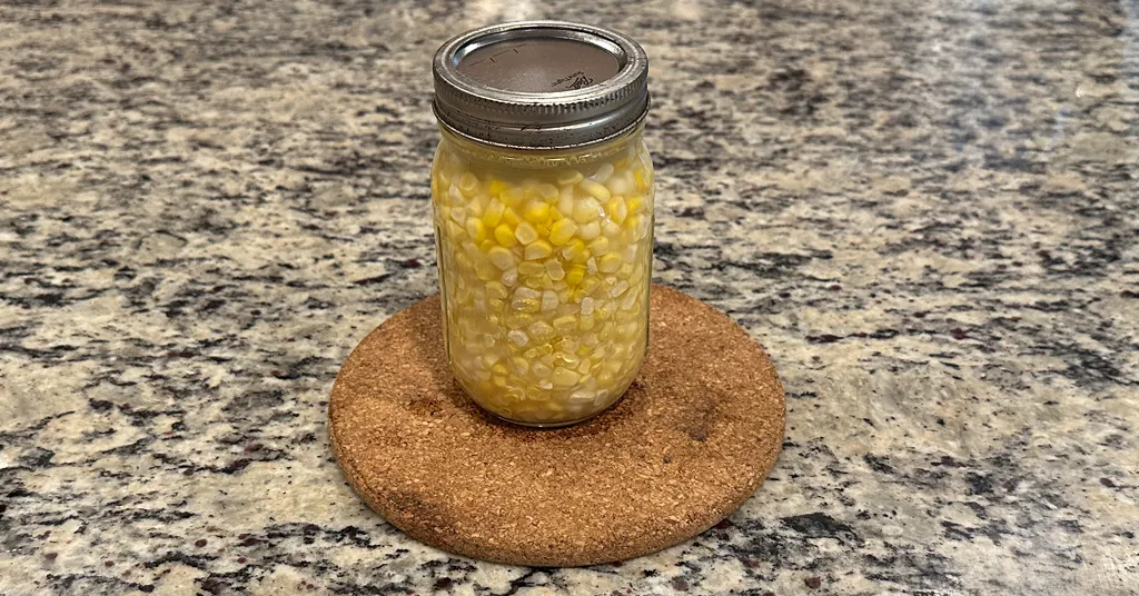 A single mason jar filled with pressure-canned corn kernels placed on a round cork trivet on a granite countertop.