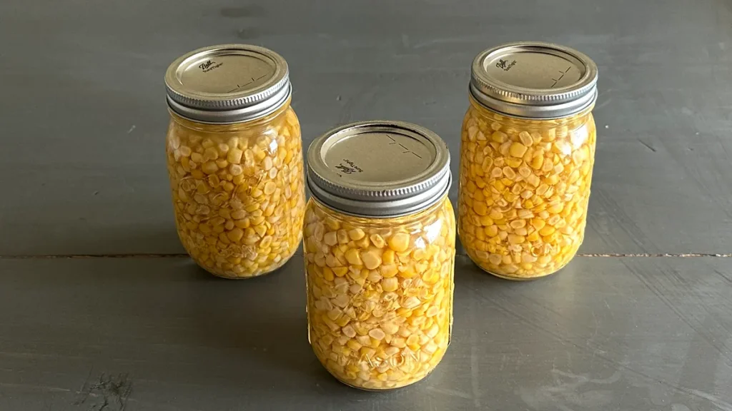 Three mason jars filled with pressure-canned corn kernels, arranged in a triangular formation on a dark wooden surface.