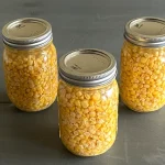 Three mason jars filled with pressure-canned corn kernels, arranged in a triangular formation on a dark wooden surface.