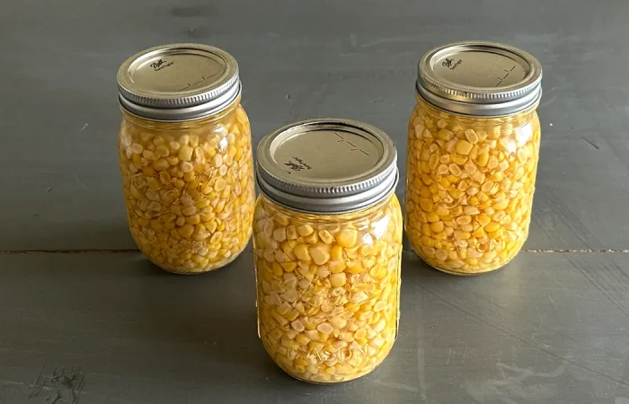 Three mason jars filled with pressure-canned corn kernels, arranged in a triangular formation on a dark wooden surface.