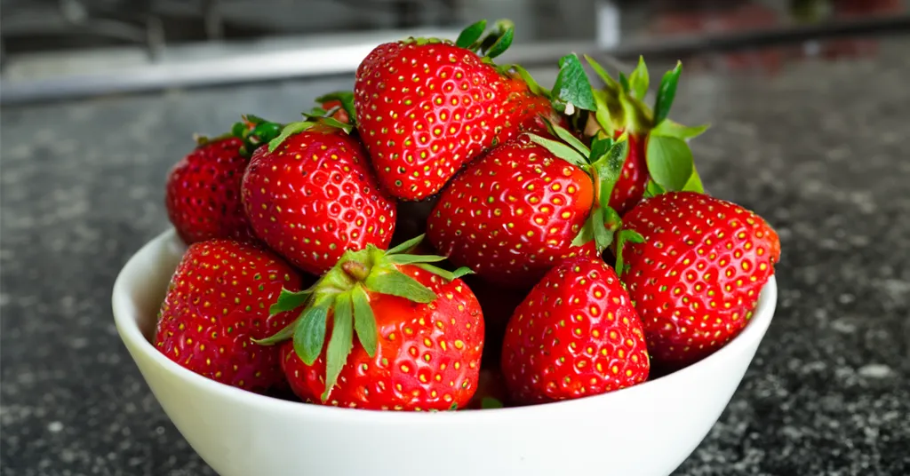 A white bowl filled with fresh, red strawberries sits on a dark countertop. The strawberries are vibrant and ripe, showcasing their bright red color and green leaves. The background is slightly blurred, drawing focus to the fruit.