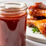 A jar of rich, dark BBQ sauce with a plate of glazed, smoky chicken drumsticks in the background on a white marble surface.