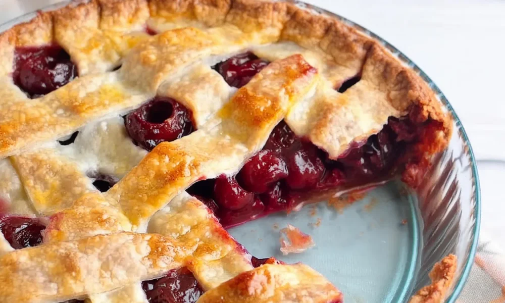 A close-up of a freshly baked cherry pie with a golden-brown lattice crust. A slice has been taken out, revealing the juicy, red cherry filling. The pie is presented in a blue pie dish on a light background.