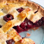 A close-up of a freshly baked cherry pie with a golden-brown lattice crust. A slice has been taken out, revealing the juicy, red cherry filling. The pie is presented in a blue pie dish on a light background.