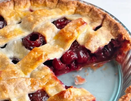 A close-up of a freshly baked cherry pie with a golden-brown lattice crust. A slice has been taken out, revealing the juicy, red cherry filling. The pie is presented in a blue pie dish on a light background.