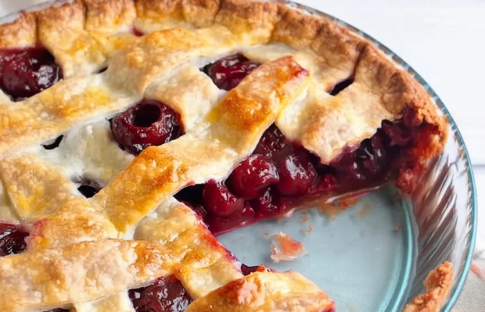 A close-up of a freshly baked cherry pie with a golden-brown lattice crust. A slice has been taken out, revealing the juicy, red cherry filling. The pie is presented in a blue pie dish on a light background.