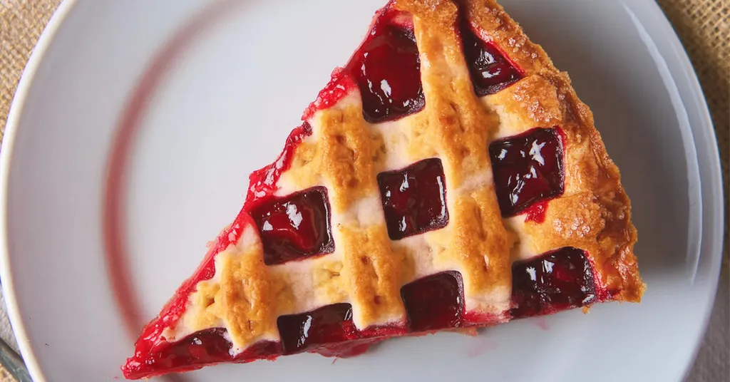 A slice of cherry pie with a golden-brown lattice crust, served on a white plate. The filling is vibrant red with visible whole cherries, contrasting with the flaky, lightly browned crust. The pie slice is neatly cut, showcasing the rich, juicy filling.