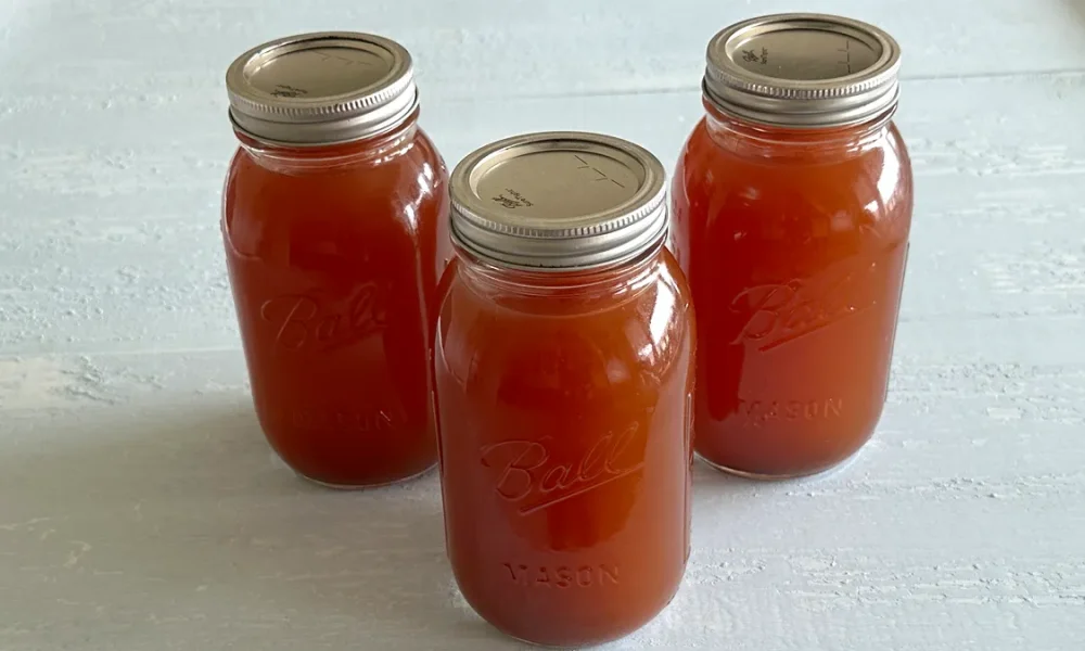 Three mason jars filled with homemade vegetable stock, sealed with metal lids, arranged on a light-colored surface. The jars are labeled "Ball Mason" and contain rich, amber-colored stock, ready for storage or use in cooking.