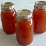 Three mason jars filled with homemade vegetable stock, sealed with metal lids, arranged on a light-colored surface. The jars are labeled "Ball Mason" and contain rich, amber-colored stock, ready for storage or use in cooking.