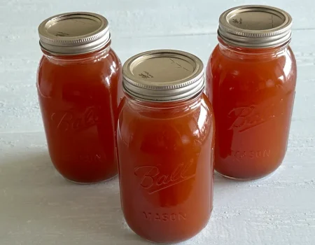 Three mason jars filled with homemade vegetable stock, sealed with metal lids, arranged on a light-colored surface. The jars are labeled "Ball Mason" and contain rich, amber-colored stock, ready for storage or use in cooking.