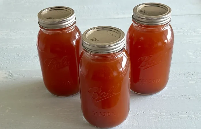 Three mason jars filled with homemade vegetable stock, sealed with metal lids, arranged on a light-colored surface. The jars are labeled "Ball Mason" and contain rich, amber-colored stock, ready for storage or use in cooking.