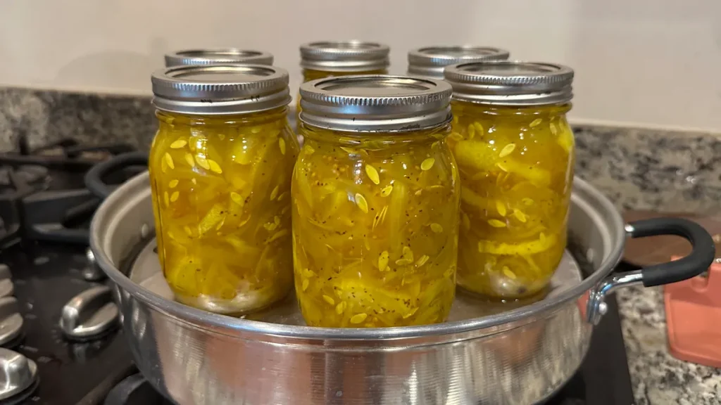 Six mason jars filled with bread and butter pickles being processed in a steam canner on a stovetop. The pickles are a vibrant yellow color, tightly packed with visible seeds and spices.