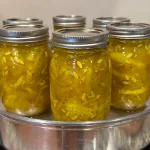 Six mason jars filled with bread and butter pickles being processed in a steam canner on a stovetop. The pickles are a vibrant yellow color, tightly packed with visible seeds and spices.