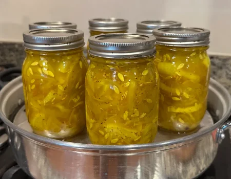 Six mason jars filled with bread and butter pickles being processed in a steam canner on a stovetop. The pickles are a vibrant yellow color, tightly packed with visible seeds and spices.