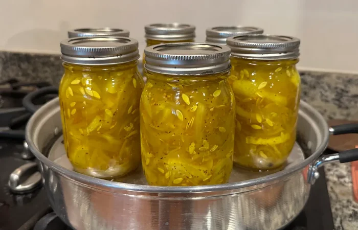 Six mason jars filled with bread and butter pickles being processed in a steam canner on a stovetop. The pickles are a vibrant yellow color, tightly packed with visible seeds and spices.