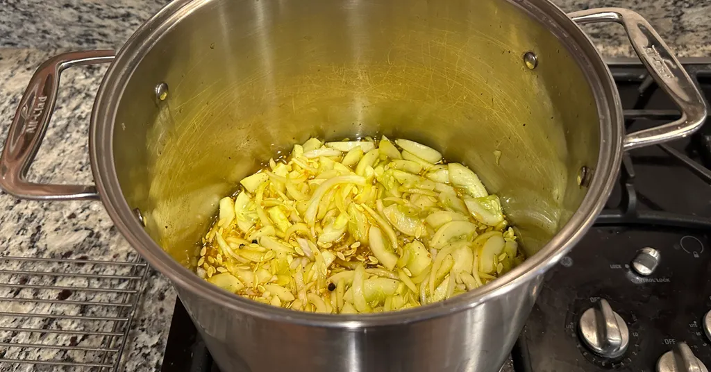 Large stainless steel pot on a stovetop filled with sliced cucumbers, onions, and spices simmering in a brine mixture, preparing bread and butter pickles.