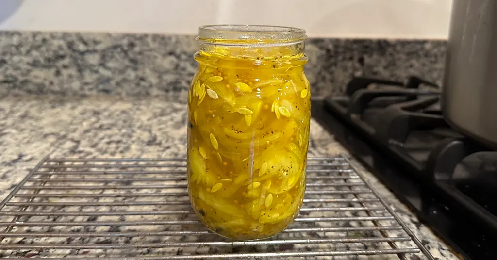 A single glass jar filled with bright yellow bread and butter pickles sitting on a wire rack after being filled, with a granite countertop and stove in the background.