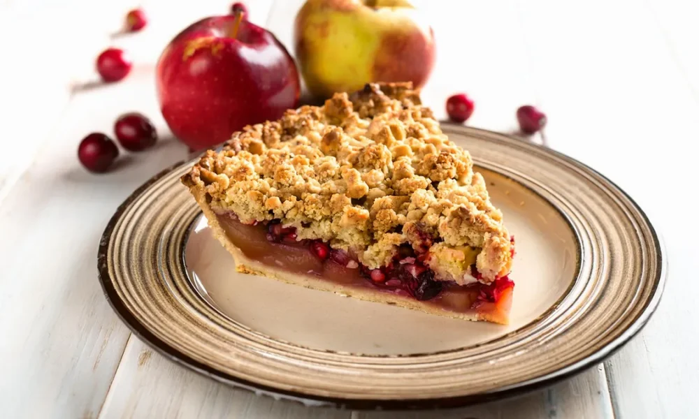 Slice of apple pear cranberry pie sitting on a plate with a streusel topping with apples and cranberries in the background.