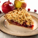 Slice of apple pear cranberry pie sitting on a plate with a streusel topping with apples and cranberries in the background.