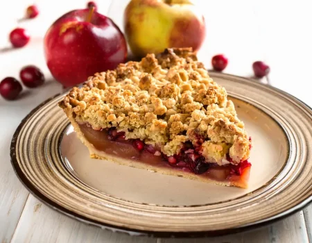 Slice of apple pear cranberry pie sitting on a plate with a streusel topping with apples and cranberries in the background.