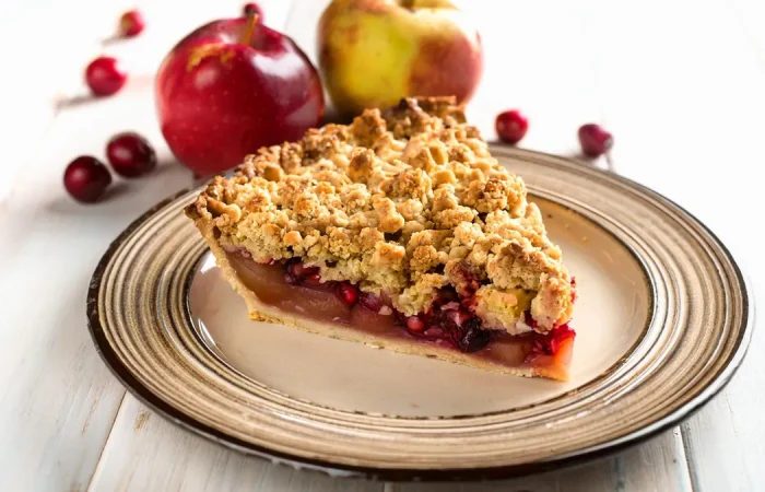 Slice of apple pear cranberry pie sitting on a plate with a streusel topping with apples and cranberries in the background.