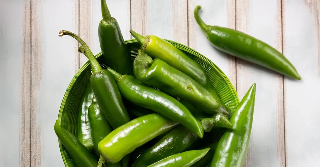 Green hatch chile peppers in a green basket sitting on a wooden surface.