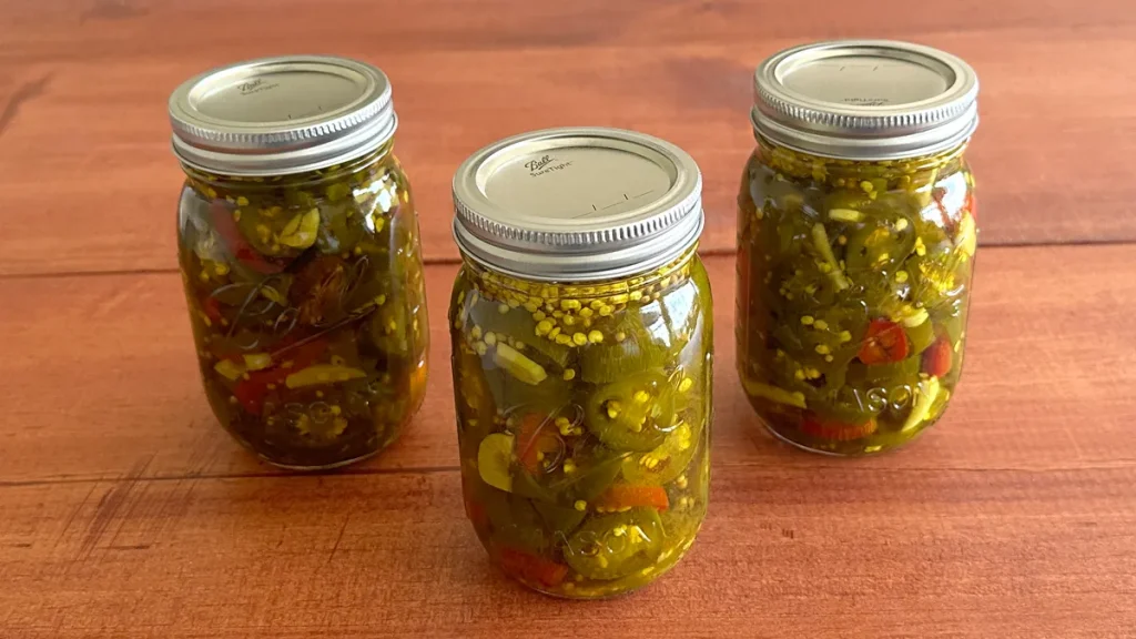 Three mason jars of Candied Jalapenos (Cowboy Candy) sitting on a wooden surface.