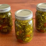 Three mason jars of Candied Jalapenos (Cowboy Candy) sitting on a wooden surface.