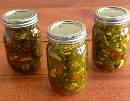 Three mason jars of Candied Jalapenos (Cowboy Candy) sitting on a wooden surface.