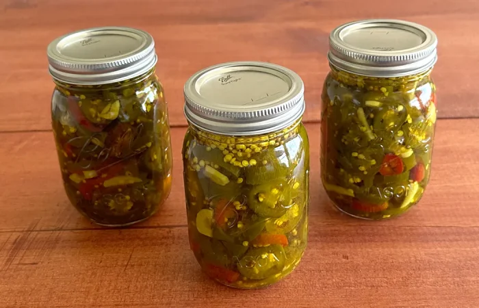 Three mason jars of Candied Jalapenos (Cowboy Candy) sitting on a wooden surface.