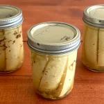 Three mason jars of dill pickle spears sitting on a wooden surface.