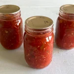 Three mason jars of fresh vegetable salsa that have been canned sitting on a pale blue surface.